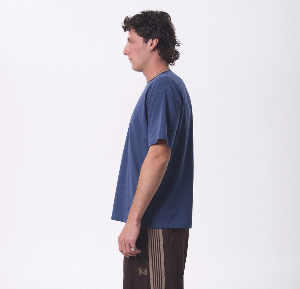 A man shown in profile wearing a loose, short-sleeve navy blue t-shirt and dark brown pants with beige stripes, standing against a plain white studio background.