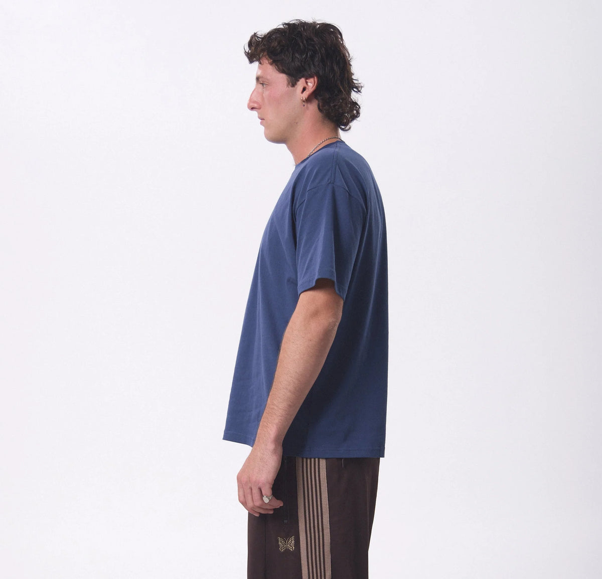 A man shown in profile wearing a loose, short-sleeve navy blue t-shirt and dark brown pants with beige stripes, standing against a plain white studio background.